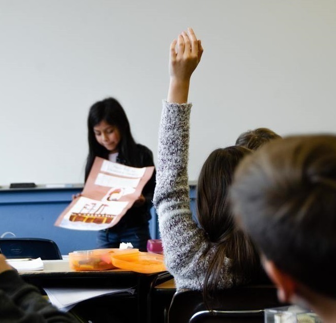 A student raising their hand while another student presents in class