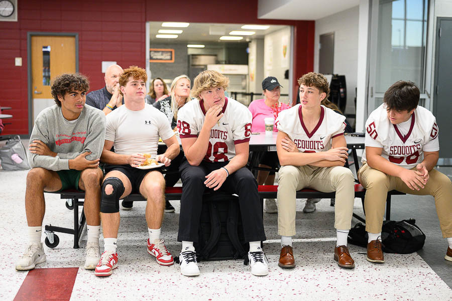 Five students sit on a bench in a school cafeteria; three wear red “SEQUOITS” football jerseys while two are in casual athletic wear, with backpacks and a food tray nearby