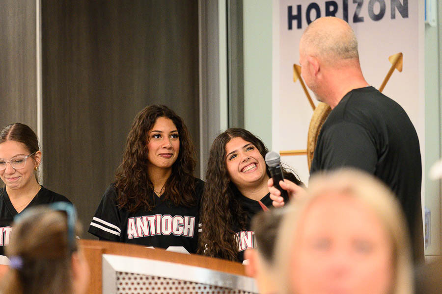 Two students wearing Antioch school apparel smiling and holding a microphone while speaking with an adult presenter on stage.