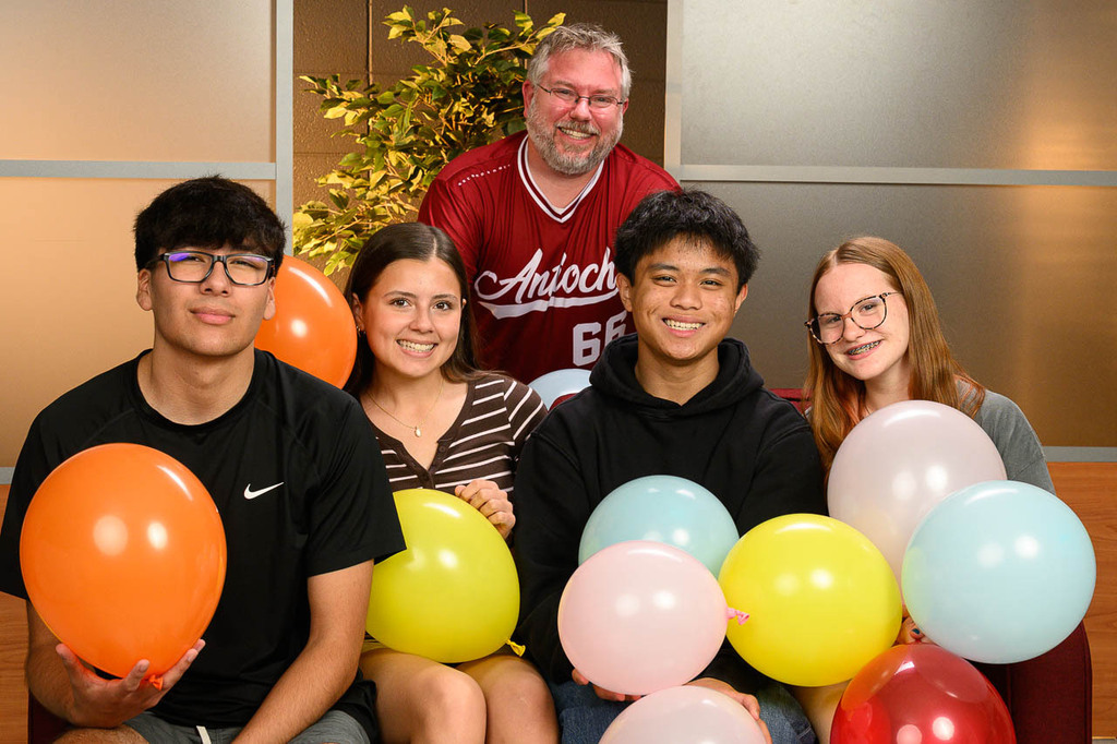 Four smiling students with balloons on couch. Teacher stands behind, smiling.