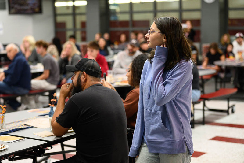 A student in a light purple hoodie standing in a crowded school cafeteria during an event.