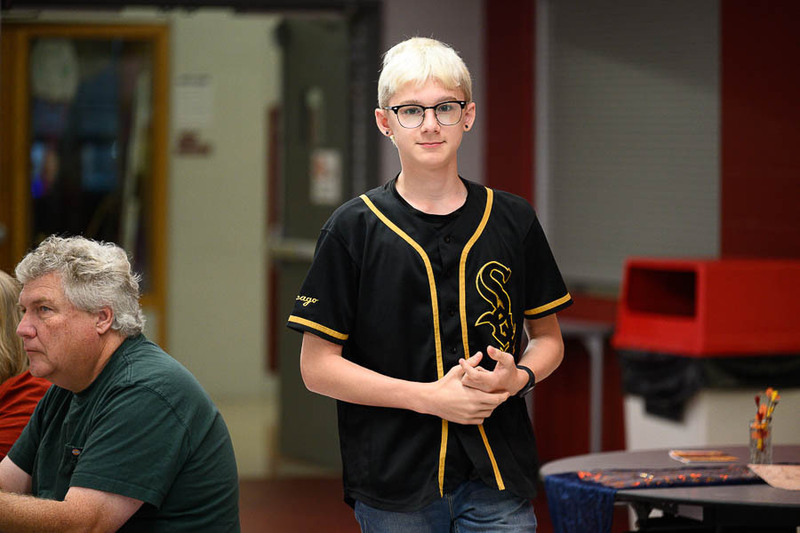 A student with blonde hair wearing a Chicago White Sox jersey standing indoors at a school event.