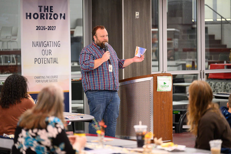 A presenter in a plaid shirt speaking into a microphone while holding a booklet on stage.
