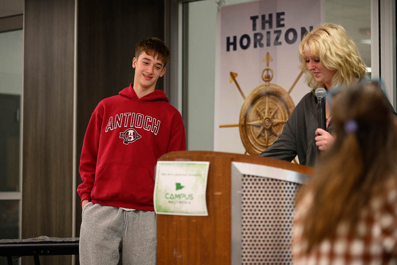 A student in a red Antioch sweatshirt standing at a podium while a presenter speaks into a microphone.