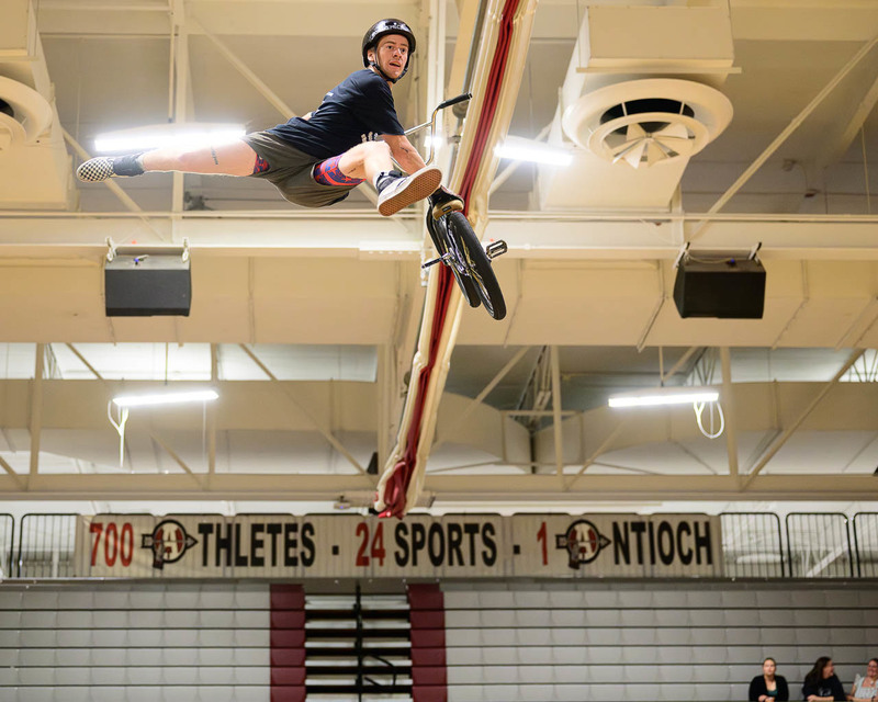 BMX rider mid‑air trick in gymnasium, one leg extended, spectators watching.