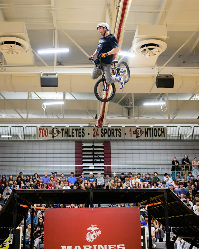  BMX rider mid‑backflip indoors, spectators in bleachers, Antioch banner visible.