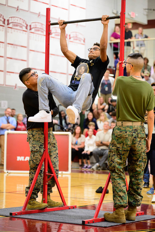  Student doing pull‑up on red bar, assisted by Marines in uniform, gym audience watching.
