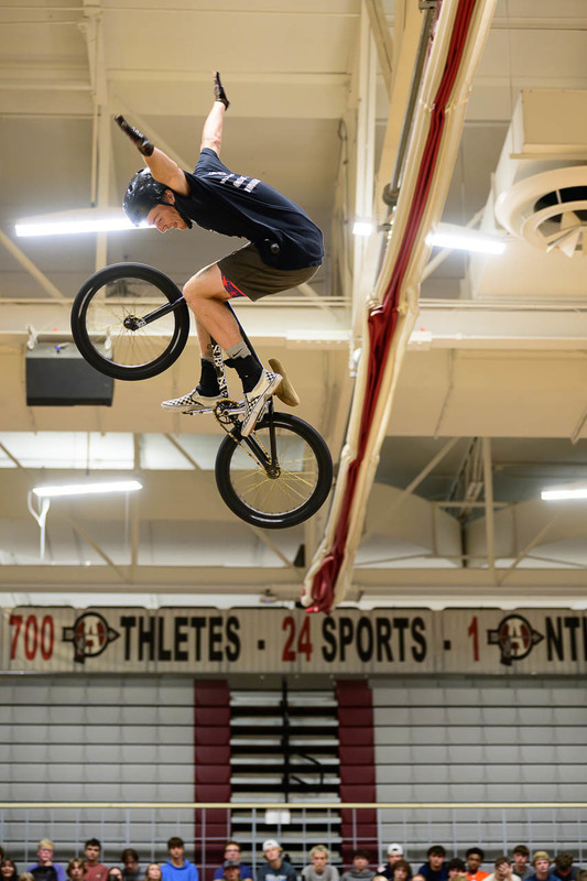 BMX rider high in air, hands off handlebars, crowd and scoreboard in gym.