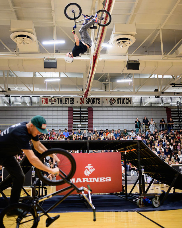 Two BMX riders performing in gym, one mid‑backflip, Marines banner on ramp, audience in bleachers.