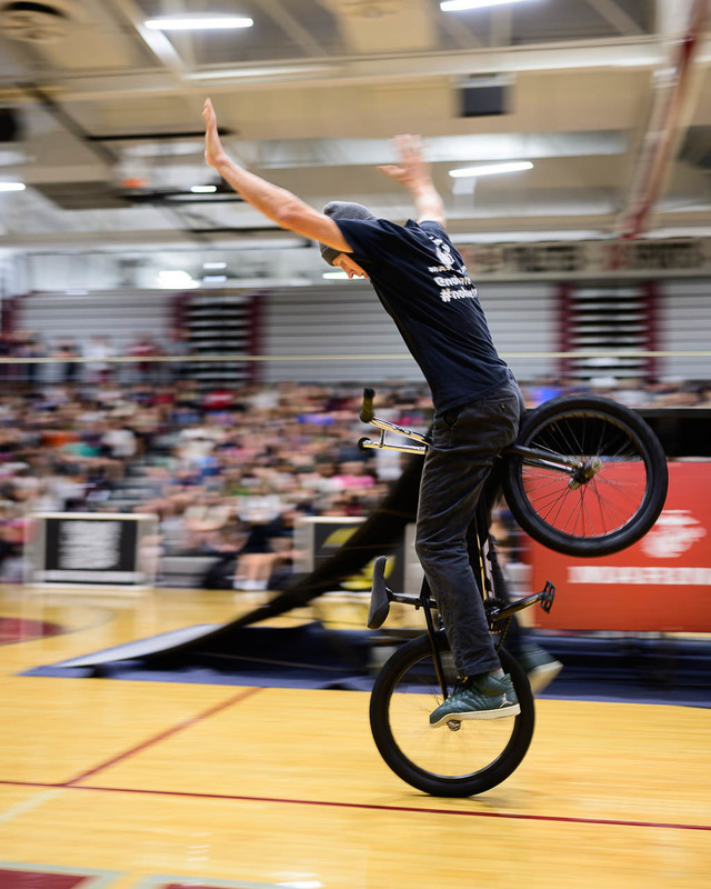 BMX rider balancing on front wheel indoors, arms raised, crowd in bleachers.