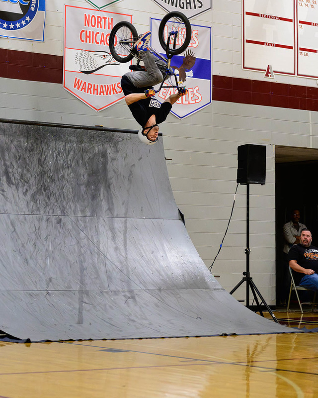 BMX rider upside down in backflip above ramp inside school gym.