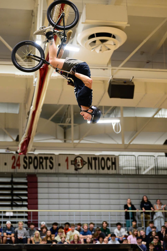 BMX rider mid‑backflip indoors, spectators in bleachers, Antioch banner visible.