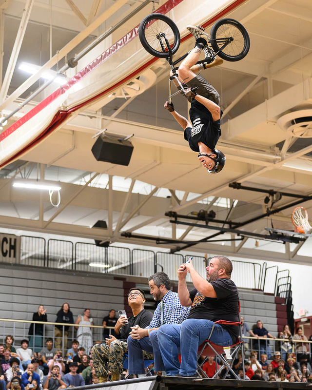 BMX rider upside down in backflip, three people below watching and filming.