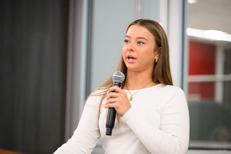 A girl in a white long-sleeve shirt speaks into a microphone in front of glass panels and a red wall, appearing engaged in a presentation or classroom talk.