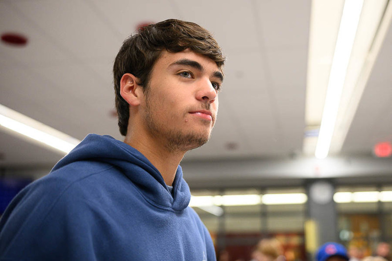 A young person in a blue hoodie stands indoors, looking slightly upward, with windows and blurred figures in the background of a school or community space.
