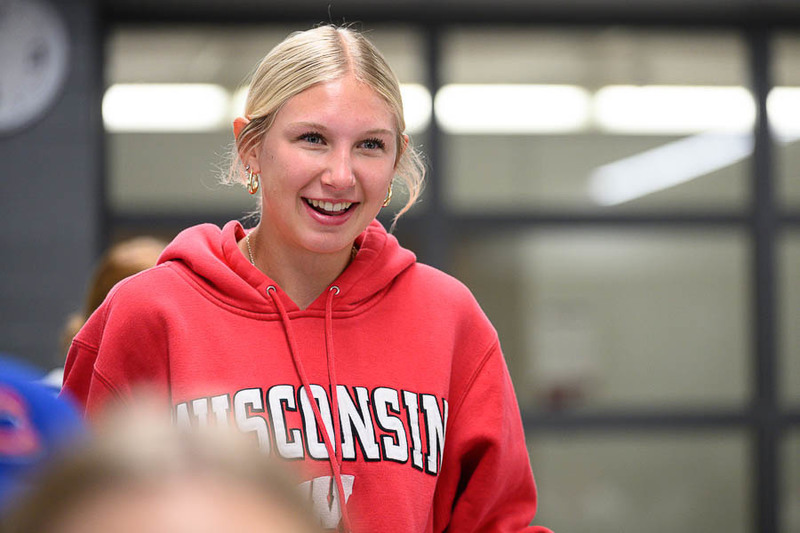 A person in a red hoodie with “WISCONSIN” printed across the front smiles indoors under fluorescent lights, with a classroom clock visible in the background.