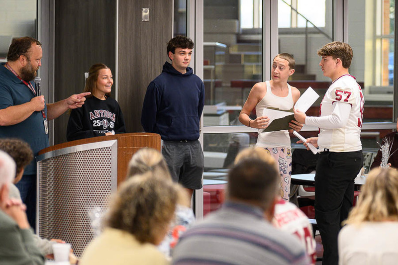 A group of students and a teacher stand near a podium in a school event; one student in an “Antioch 24-25” sweatshirt smiles while others listen and interact.