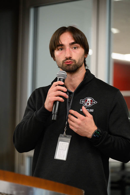 A person in a black jacket with a “WRESTLING” logo speaks into a microphone, wearing a lanyard and smartwatch, in a modern indoor setting with glass panels.