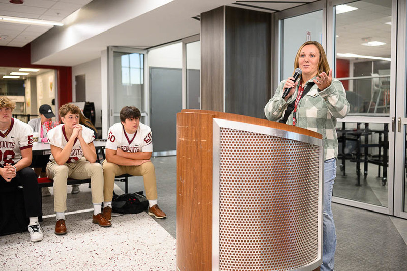 A woman in a plaid jacket speaks into a microphone at a podium in a school setting, with four football players in matching Sequoits jerseys seated beside her.
