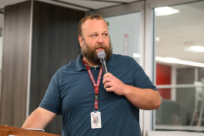 A man in a dark blue polo shirt and red lanyard speaks at a podium with a microphone, in a modern indoor space with glass walls.