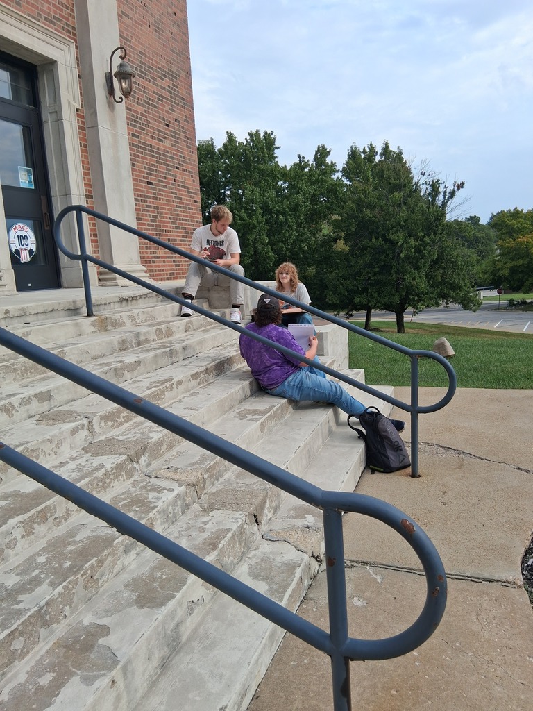 Students sit on steps outside a building creating artwork.