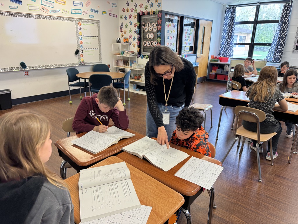 A teacher helps a student sitting at a desk in the classroom think through a math equation in a workbook.
