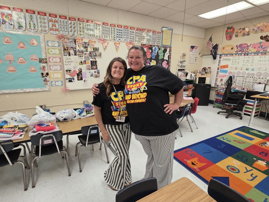 Two people stand in a classroom with tables, chairs, and a colorful rug.