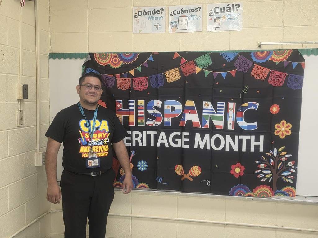 A man stands before a banner celebrating Hispanic Heritage Month, featuring vibrant designs and colorful text.