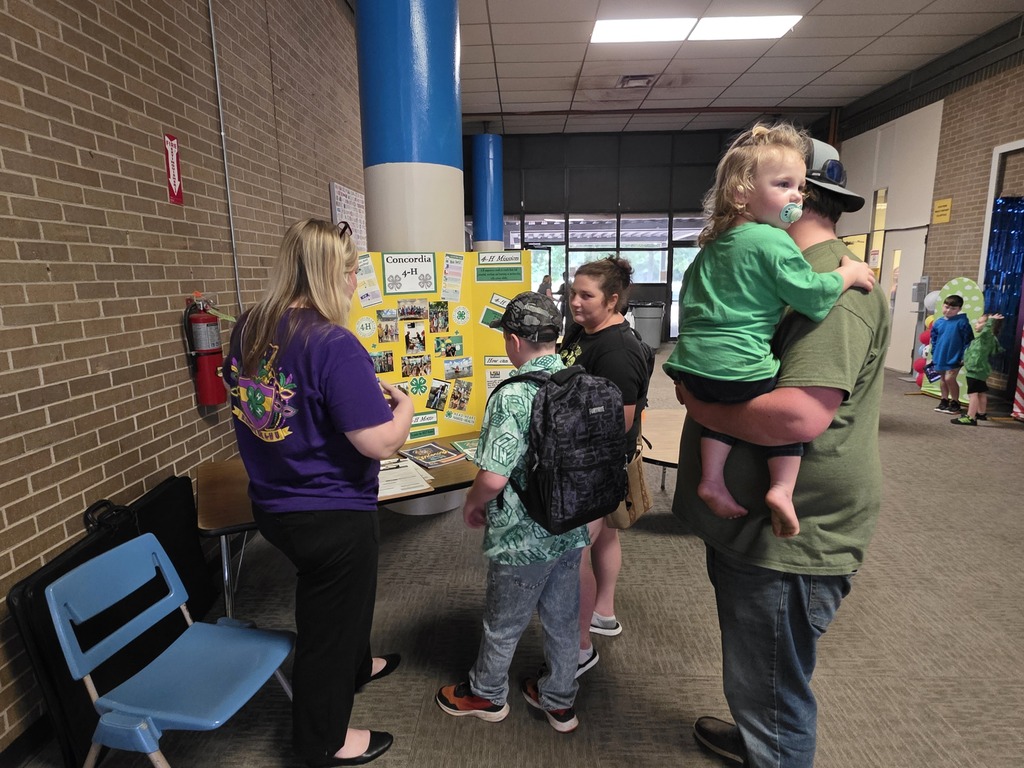 A small group of adults and children, some carrying children, examine a bulletin board in a hallway.