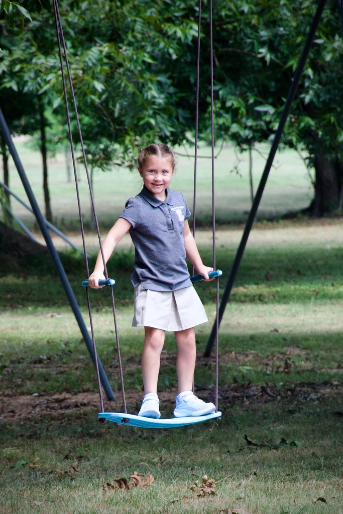 Child wearing a blue shirt and skirt standing on a blue swing suspended from ropes in a park.
