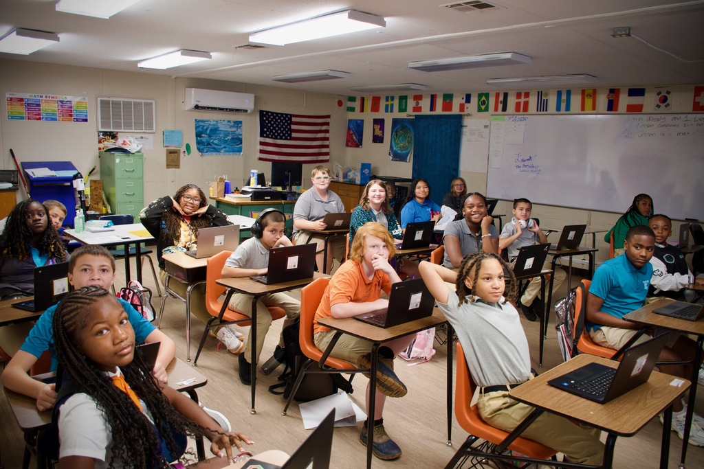 A classroom with several students at desks, using laptops. A whiteboard is on the wall, and various flags hang.