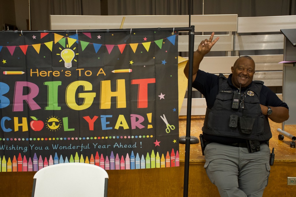 A man in a police uniform smiles while standing next to a sign that reads "Here's To A Bright School Year!"