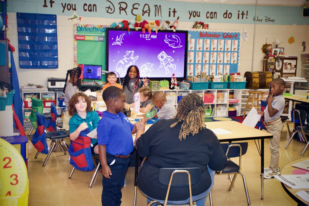 Classroom with students seated at tables. A woman leads a discussion. A large monitor on the wall displays an image.
