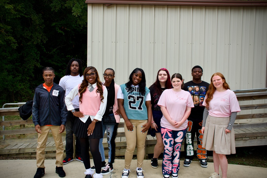 A group of teens stands in front of a building with benches and trees behind them.