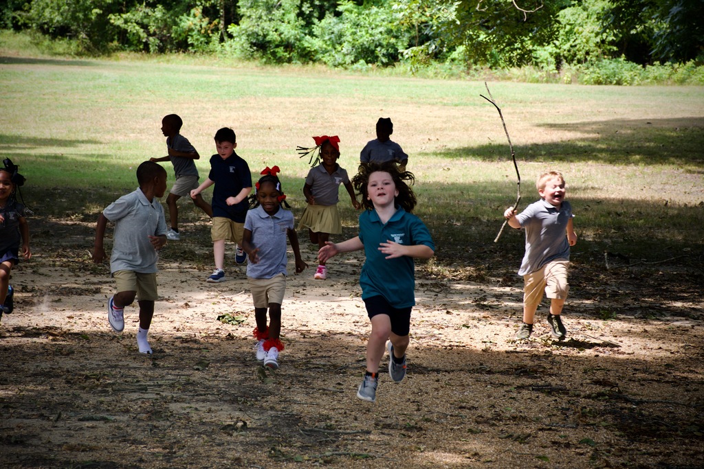 Several children run outdoors in a shaded area with a dirt path, some carrying sticks, and a green grassy field in the background.