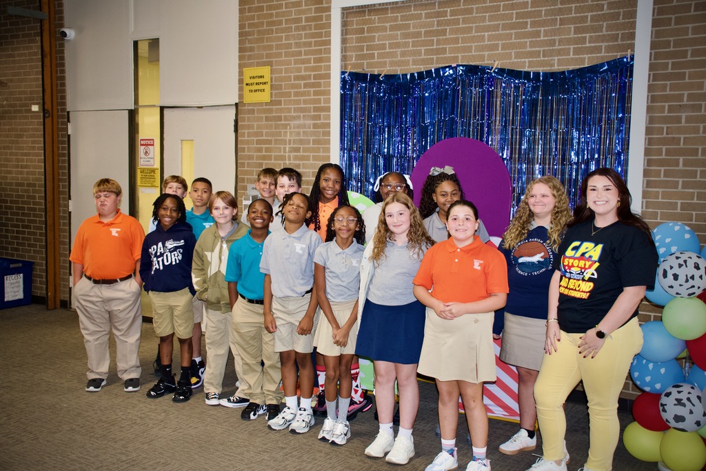 A group of children and adults in uniforms stand together in front of a blue backdrop with balloons.