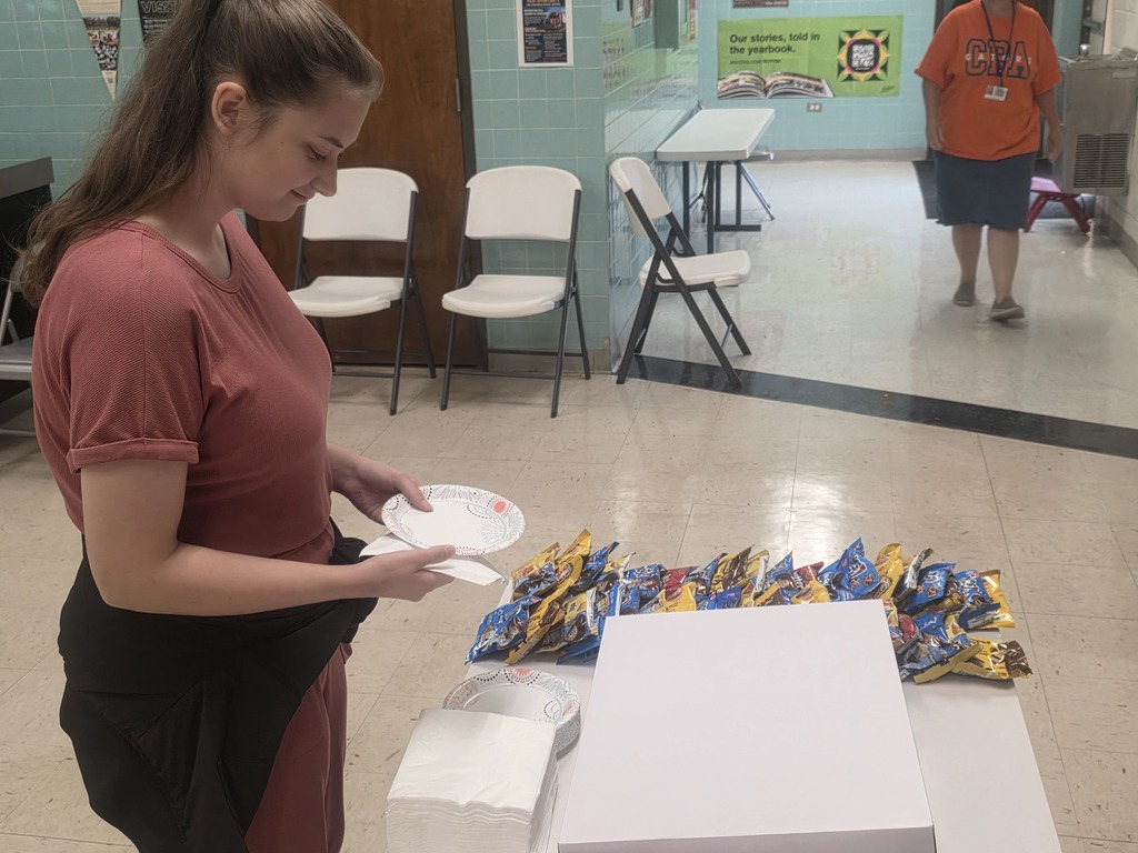 Woman at a table holds a plate and bag of chips. Behind her, another woman in an orange shirt. Chairs and a table in the background.