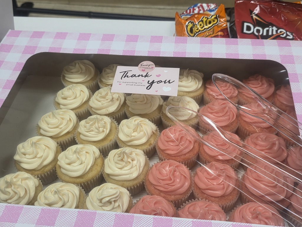 A box of cupcakes with pink frosting, some with white frosting. A note reads "Thank you". Background has snacks.