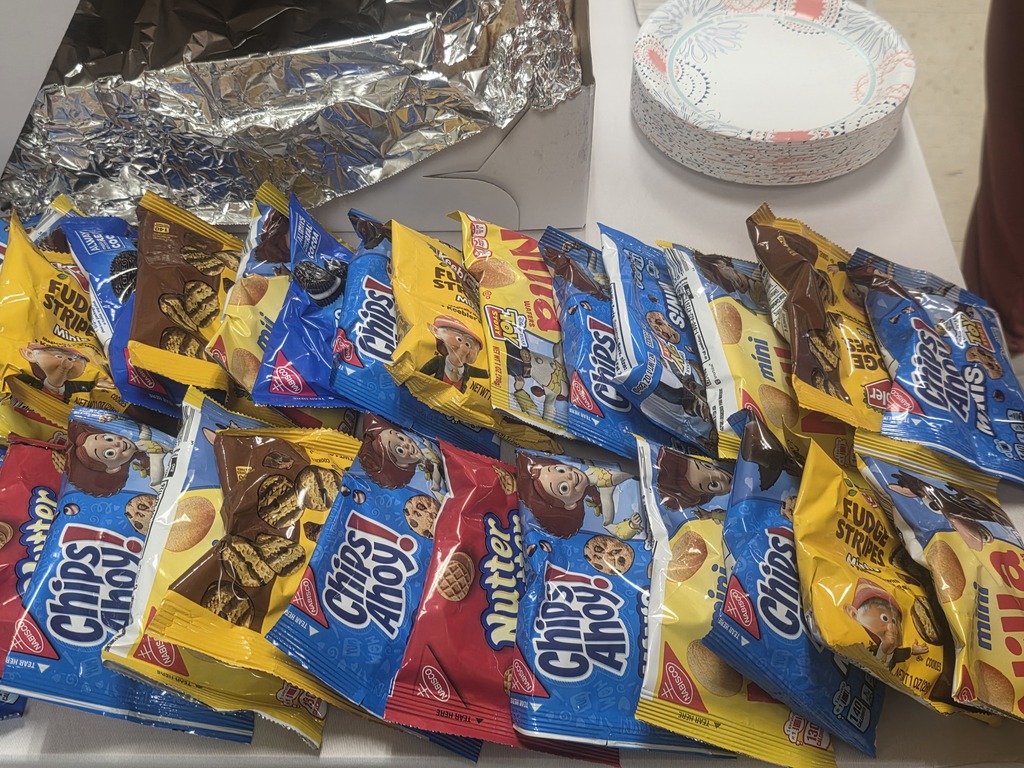 A table holds numerous bags of chips. One brand has blue packaging, another has yellow, and some have cartoon characters on them.