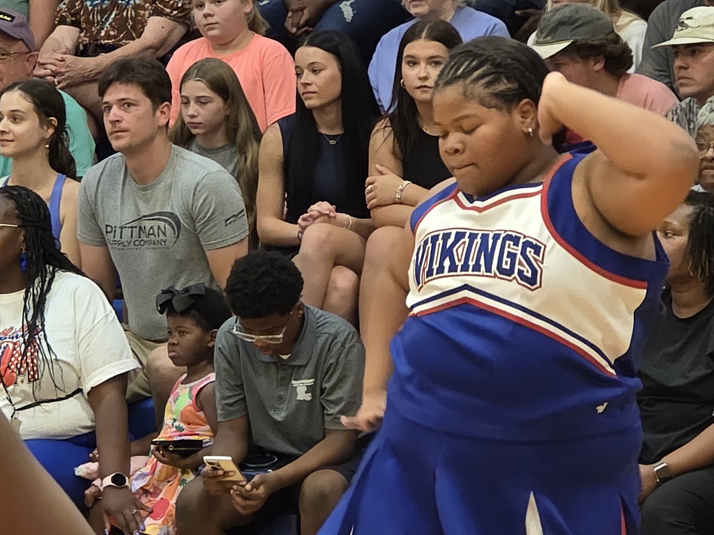 A group of people sitting in bleachers; a young girl in a Vikings cheerleader uniform is standing.