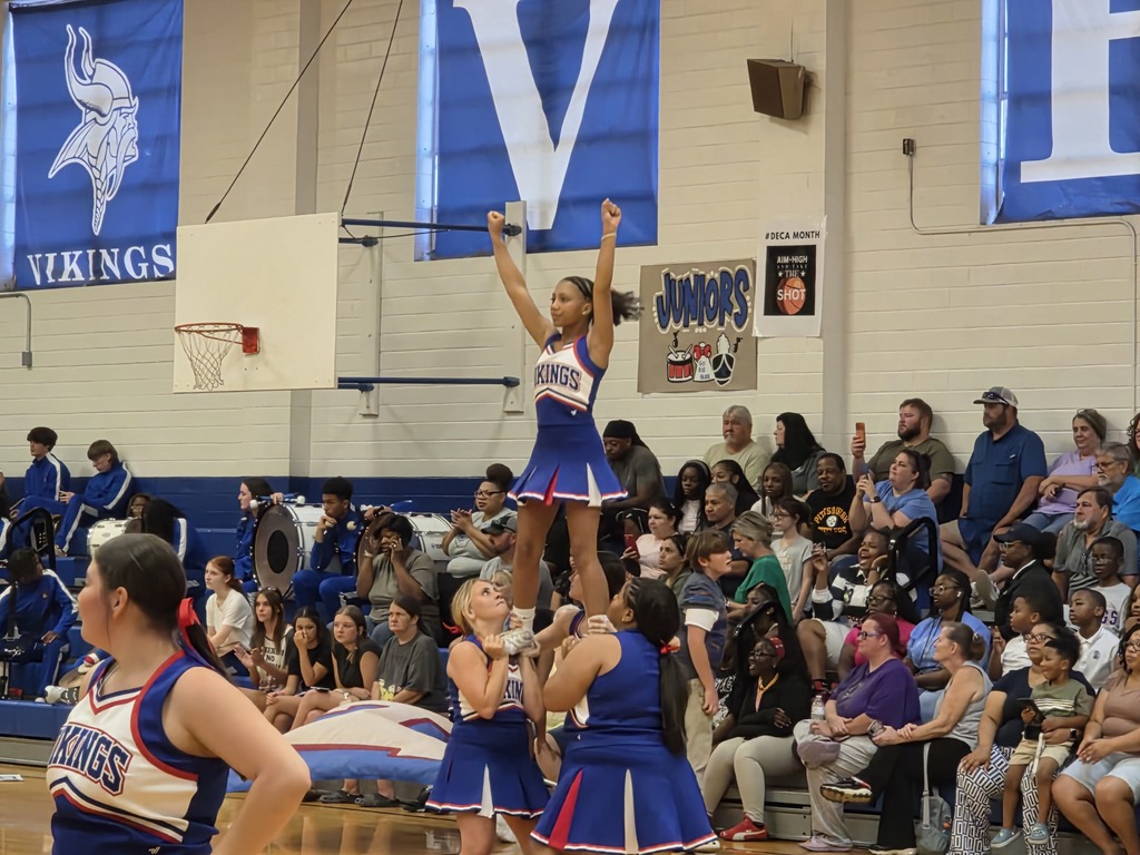 Cheerleaders perform stunts on a basketball court; one cheerleader is on shoulders of two others.