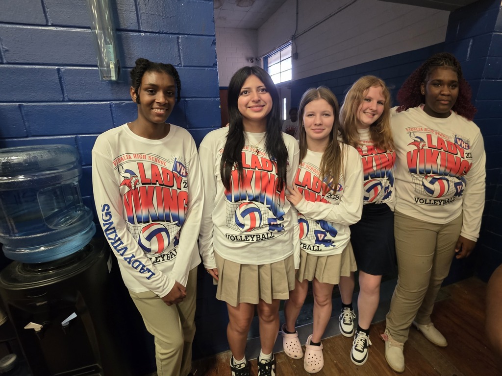 Five women wearing matching shirts are in a room with blue walls. They stand next to a water cooler.