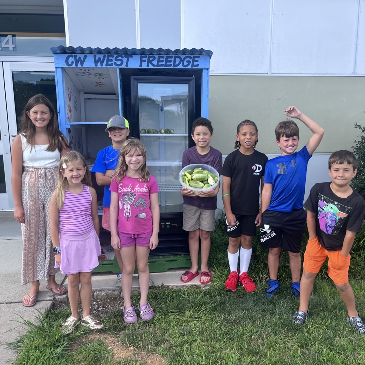 students standing with a bowl of cucumbers they picked. There is a fridge behind them.