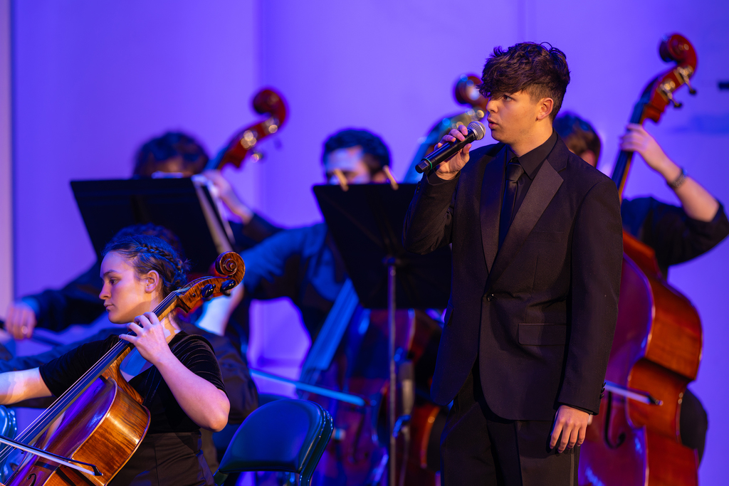 A student in a black suit sings while the orchestra plays in the background.