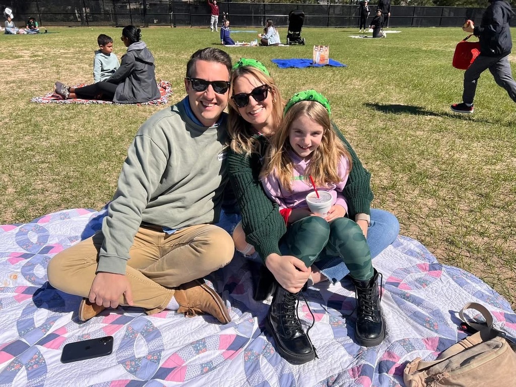 Three people sitting closely on a patterned blanket in a sunny park, enjoying a casual outdoor moment together.