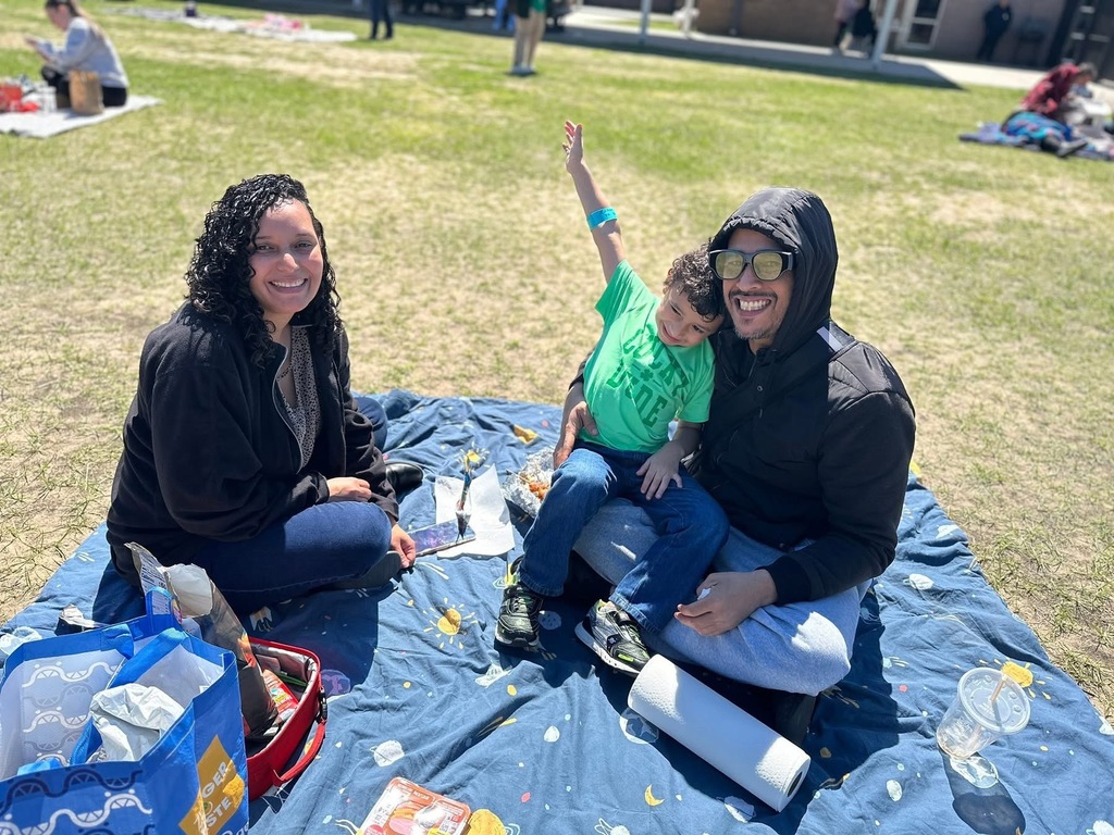 Three people sitting on a blue blanket having a picnic on a grassy field under sunny weather.