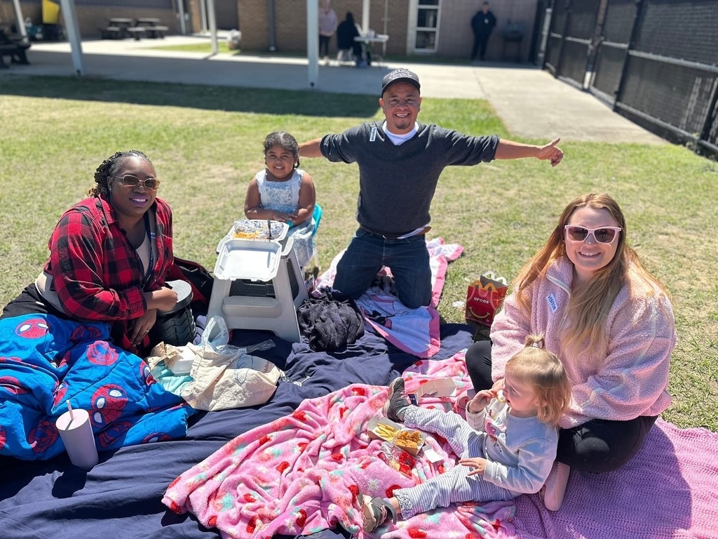 Group of five people enjoying a sunny picnic on colorful blankets in a grassy outdoor area near buildings.