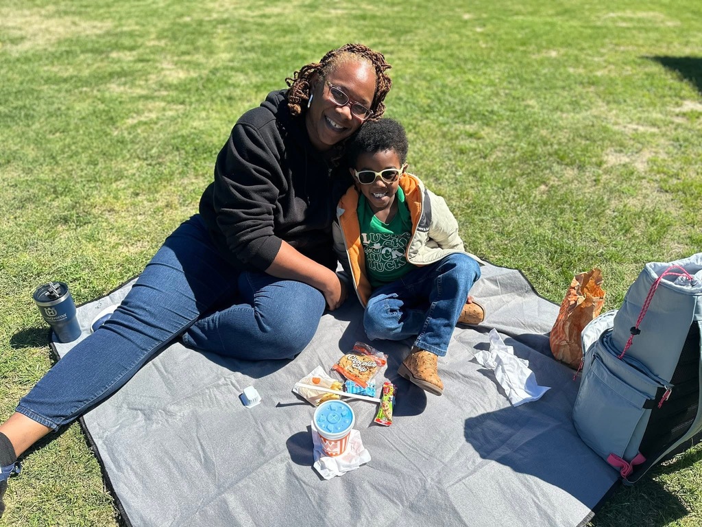 A woman and child sitting on a gray picnic blanket on green grass, enjoying snacks and drinks on a sunny day.