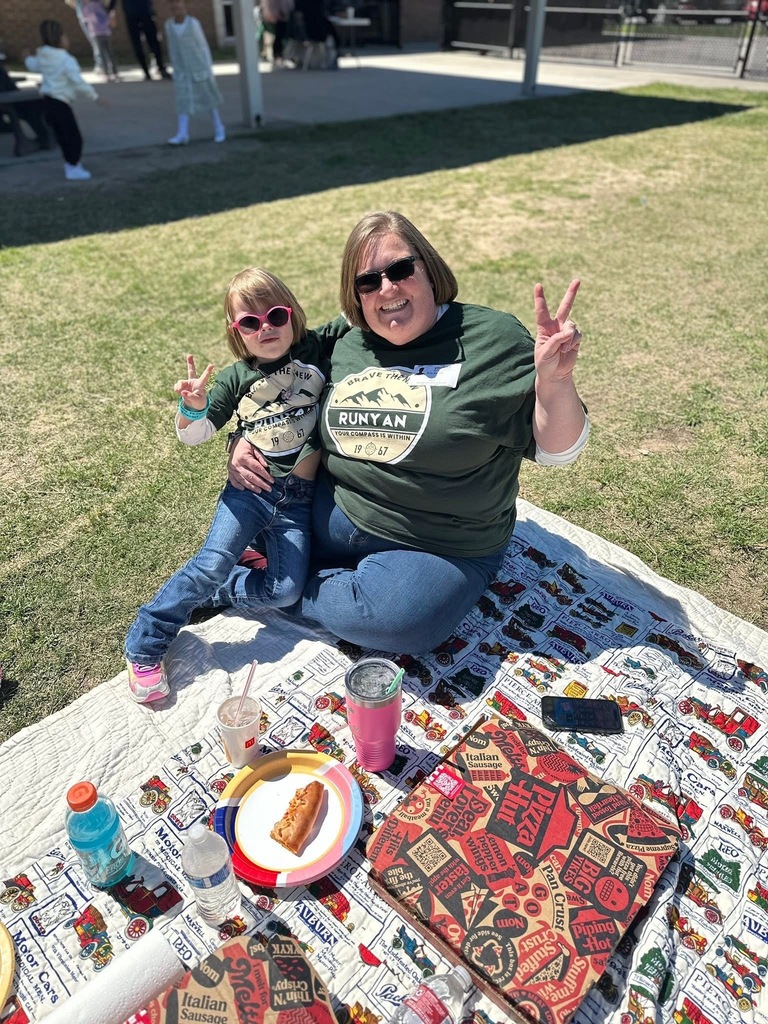 Mother and daughter sitting on a blanket with pizza box and drinks, both showing peace signs.
