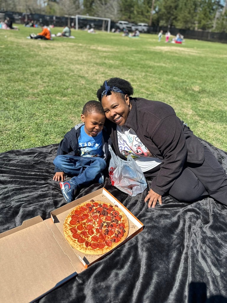 Mother and child enjoying a picnic on a grassy field with a freshly opened pepperoni pizza placed on blanket beside them ready to eat.